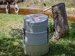 Automated water sampler labeled 'ISCO' placed on grassy ground near a stream. Tubing extends from the device toward the water. In the background, two researchers in waders are standing in the stream, slightly out of focus, conducting fieldwork. A large tree stump sits nearby on the right side.